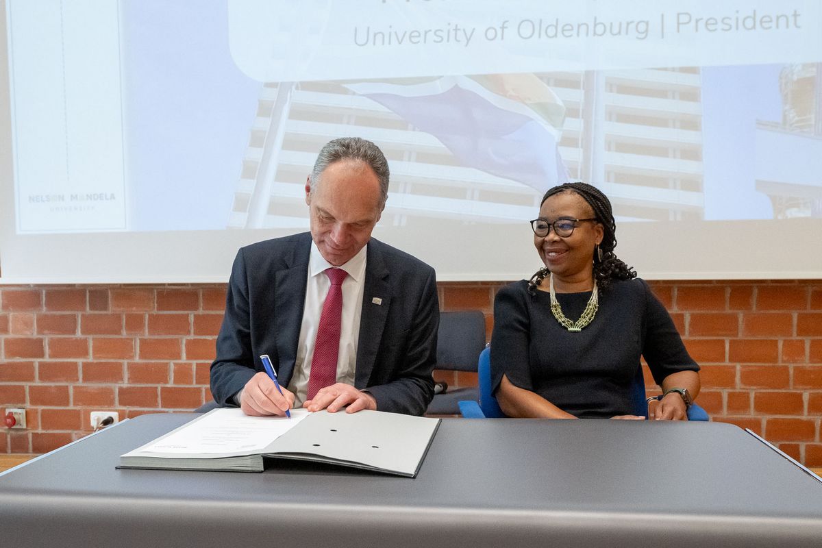 Das Bild zeigt zwei Personen, Vice-Chancellor Sibongile Muthwa (NMU) und Präsident Ralph Bruder (UOL), die an einem Tisch sitzen und ein Dokument unterzeichnen. Der Präsident der Universität Oldenburg schreibt mit einem Stift auf das Dokument. Die Vice-Chancellor der Nelson Mandela University lächelt und blickt auf das Dokument. Im Hintergrund ist ein Banner mit der Aufschrift "University of Oldenburg | President" und ein Bild des Universitätsgebäudes der NMU zu sehen. Das Bild dokumentiert die Unterzeichnung einer Vereinbarung im Rahmen des 25-jährigen Kooperationsjubiläums zwischen den beiden Universitäten.
