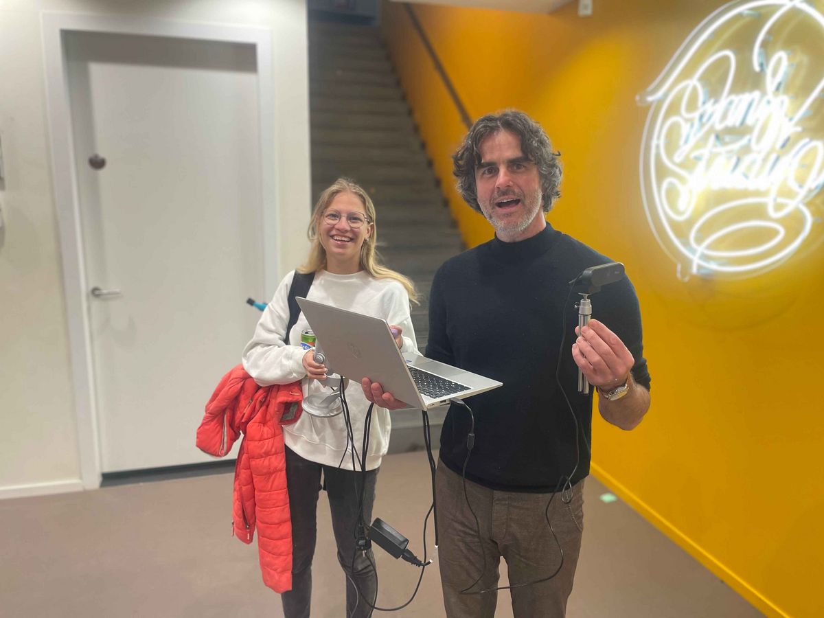 Two people under a staircase, the man with a laptop and microphone, in the background is a yellow wall with a neon installation.
