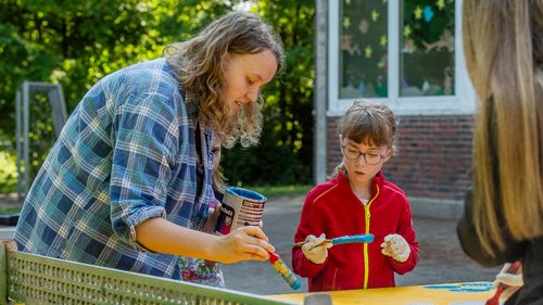A student and two girls stand around a table tennis table painted yellow, all holding brushes and painting the playing fields white on the table.