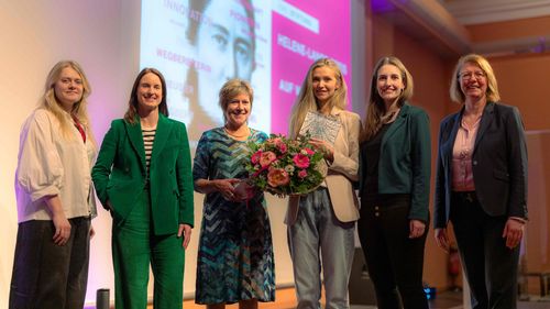 The picture shows six women standing in front of a screen onto which the logo of the Helene Lange Prize is projected. They smile into the camera. Award winner Sandra Drolshagen holds a bouquet of flowers and a certificate in her hands.