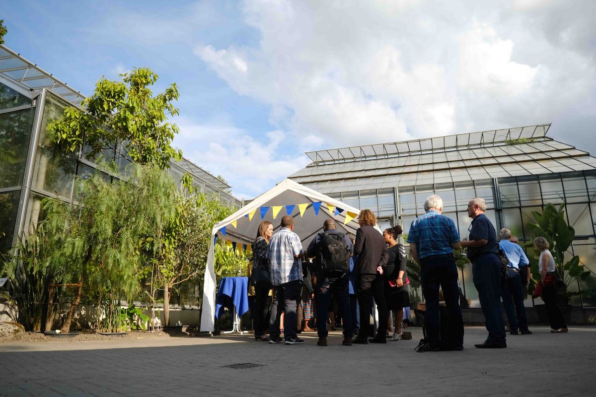 Das Bild zeigt eine Gruppe von Menschen, die sich unter einem Zelt mit bunten Fahnen versammelt haben. Das Zelt steht vor einem großen Gewächshaus mit Glasfassade. Die Personen scheinen sich zu unterhalten oder etwas zu essen und zu trinken. Die Szene vermittelt einen Eindruck von informellem Austausch und Geselligkeit im Rahmen des Welcome Dinners zum 25-jährigen Kooperationsjubiläum der Universität Oldenburg mit der Nelson Mandela University.