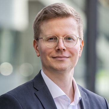 Mann mit kurzen blonden Haaren, runde Brille mit durchsichtigem Rahmen, weißes Hemd, schwarzes Jacke, lächelt in die Kamera.