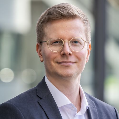 Mann mit kurzen blonden Haaren, runde Brille mit durchsichtigem Rahmen, weißes Hemd, schwarzes Jacke, lächelt in die Kamera.