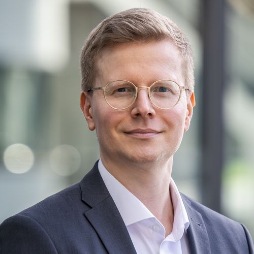 A man with short blond hair, round glasses with clear frames, a white shirt and a black jacket is smiling at the camera.