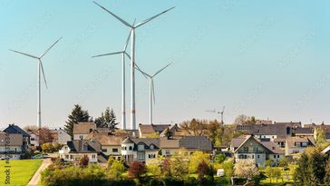 Das Bild zeigt eine Gruppe von Windrädern. Im Vordergrund befindet sich ein Dorf. Die Windräder sind groß und überragen die Szenerie. Der Himmel ist blau.