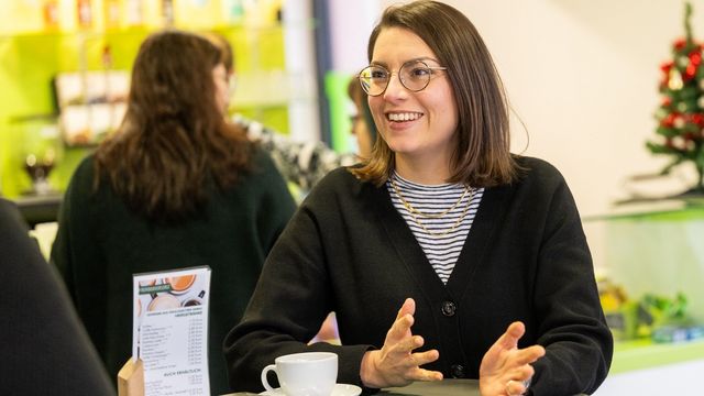 Das Bild zeigt Ann Kathrin Schubert. Sie steht in einem Café an der Uni, im Hintergrund ist die Bar zu erkennen. Sie gestikuliert leicht mit ihren Händen. Sie schaut zu der Interviewerin und lächelt diese an.