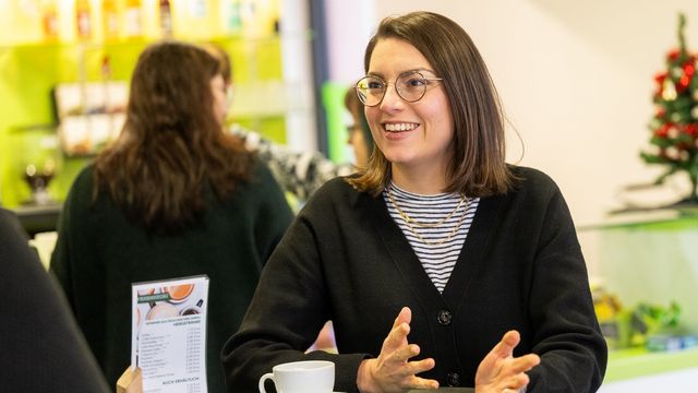 The picture shows Ann Kathrin Schubert. She is standing in a university café, with the bar in the background. She is gesturing slightly with her hands. She looks at the interviewer and smiles.