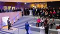 View into the interior of the HIFMB, some people are standing at bar tables, others are sitting on the steps, most are standing on a kind of gallery. One man stands somewhat apart from the guests on a ledge behind a microphone.