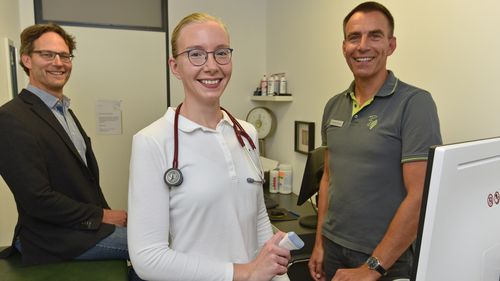 The picture shows Michael Freitag, Rike Geiken and Markus Ennen in a doctor's surgery. Rike Geiken is standing in the centre with a stethoscope around her neck and a transducer in her hand.