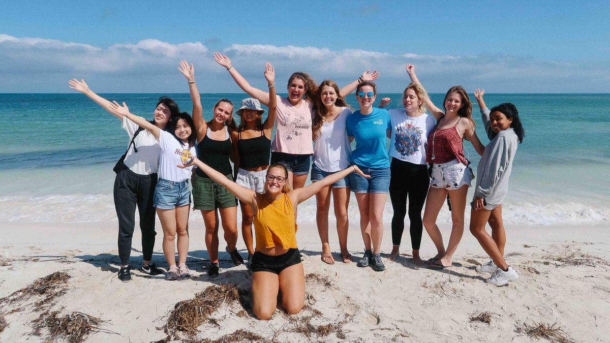 Eine Gruppe junger Frauen posiert lächelnd am Strand vor türkisfarbenem Meer und blauem Himmel.