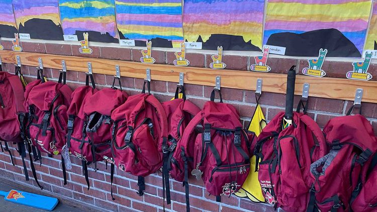 A classroom or hallway in a school decorated with children's paintings of mountains and a sunset. Red school bags with llamas and children's names hang underneath.