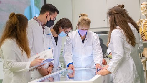Several students in white coats stand around the virtual dissection table (a large, horizontal screen). Prof. Bräuer shows something to the bystanders. 