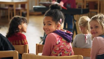 A girl of primary school age with a cochlear implant sits with her peers in a school-like situation. Everyone is sitting with their backs to the photographer, the girl with the CI turns round to face the viewer.