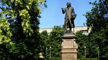 The picture shows a monument to Immanuel Kant. Kant is standing on a pedestal and looking into the distance. A park and buildings can be seen in the background.