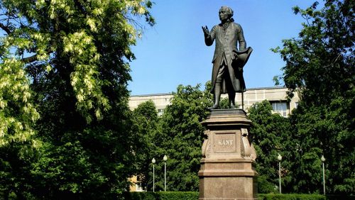 Das Bild zeigt ein Denkmal für Immanuel Kant. Kant steht auf einem Sockel und blickt in die Ferne. Im Hintergrund sind ein Park und Gebäude zu erkennen.