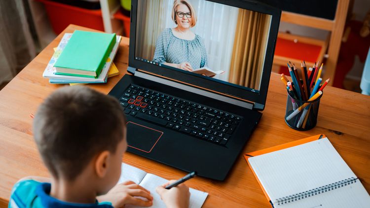 Ein Junge sitzt mit seinem Heft vor einem Laptop. Auf dem Bildschirm ist eine Lehrerin zu sehen.