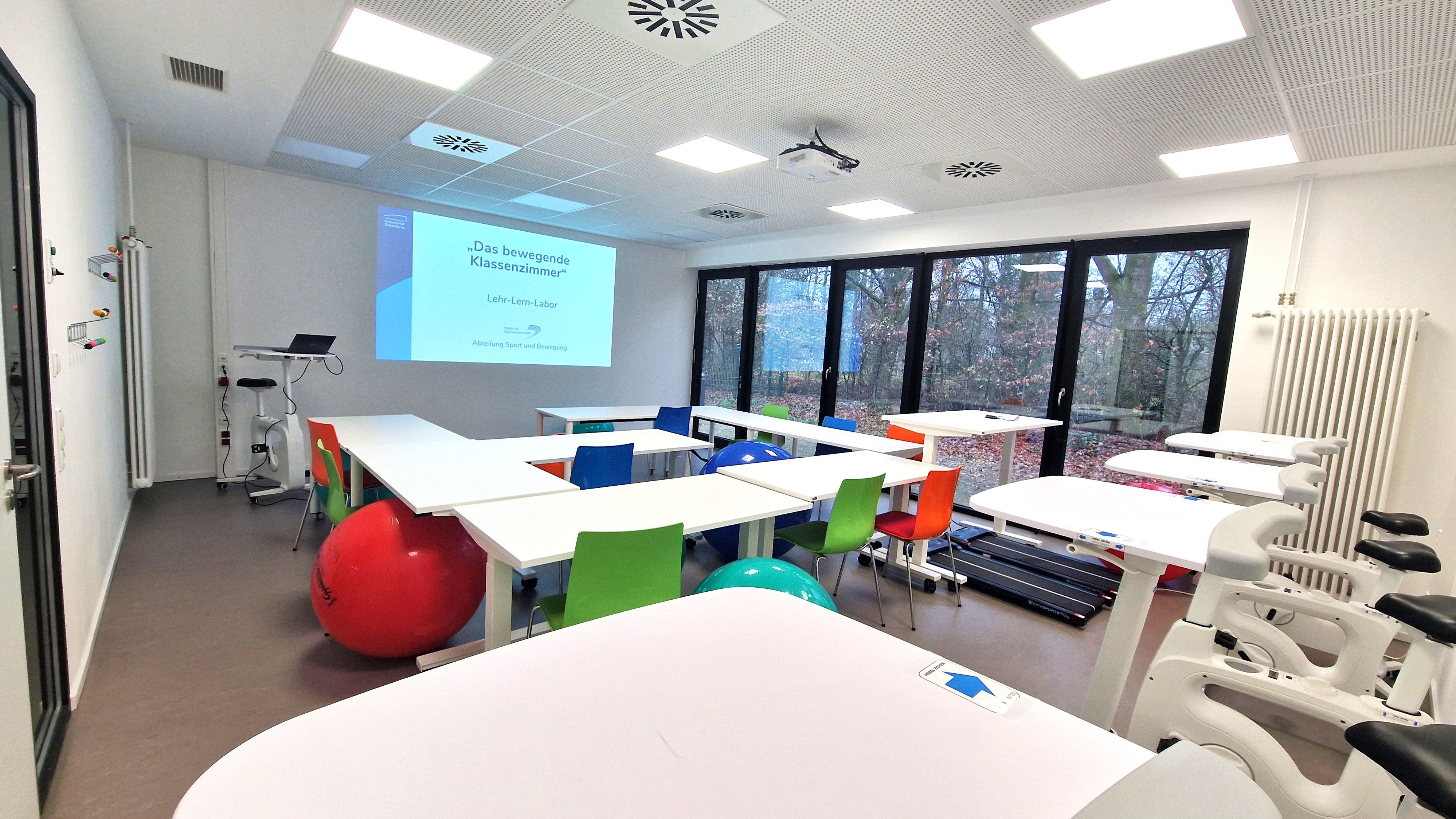 View of the seminar room from the rear left corner of the room. At the front, a presentation slide is projected onto the wall by a projector. At the back of the wall are five desk bikes next to each other, with a few tables in front of them, alternating with Pezzi balls and chairs. One table is raised, and two treadmills are positioned side by side on the floor below.