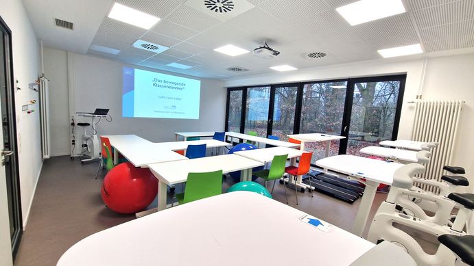 Blick von der hinteren linken Raumecke in den Seminarraum. Vorne ist eine Präsentationsfolie von einem Beamer an die Wand projiziert. Hinten an der Wand stehen fünf Deskbikes nebeneinander, davor einige Tische, an denen sich Pezzibälle und Stühle abwechseln. EIn Tisch ist hochgefahren, am Boden darunter befinden sich nebeneinander zwei Laufbänder. 