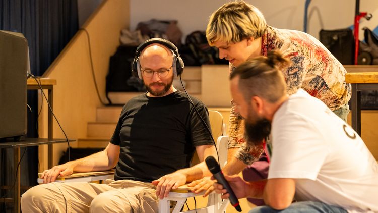 Eine Person mit Kopfhörern sitzt auf auf einem Stuhl, dem Vibro-Chair. Ein Wissenschaftler erklärt der sitzenden Person etwas. Der Moderator hockt daneben.
