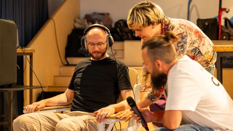 A person with headphones is sitting on the Vibro-Chair, with a scientist to the right, explaining the device. The event host is sitting next to them.