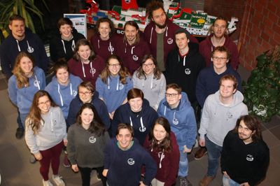 The photo shows the Environmental Sciences Student Council for winter & summer semester 2017/18. 22 students can be seen, all in their early to late 20s, all wearing colourful jumpers or jackets with the Student Council logo.  They are standing in several rows. The photo was taken in the ICBM. In the background, there is a model ship in a display case in front of a brick wall, with a potted plant to the right and left.