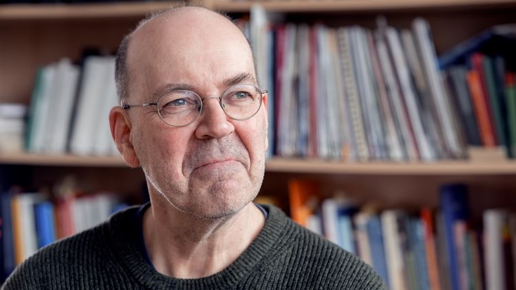 The picture shows Dietmar von Reeken. He is sitting in front of a bookshelf and looking thoughtfully out of the window.