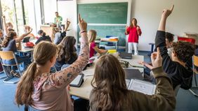 Das Bild zeigt eine Schulklasse. Vorne steht eine Lehrerin und erklärt den Schülerinnen und Schülern etwas. Hinter ihr befindet sich eine Kreidetafel. Einige der Schülerinnen und Schüler melden sich.