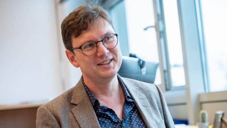 Mark Schweda in conversation. He wears glasses, a light brown jacket and a blue, patterned shirt.