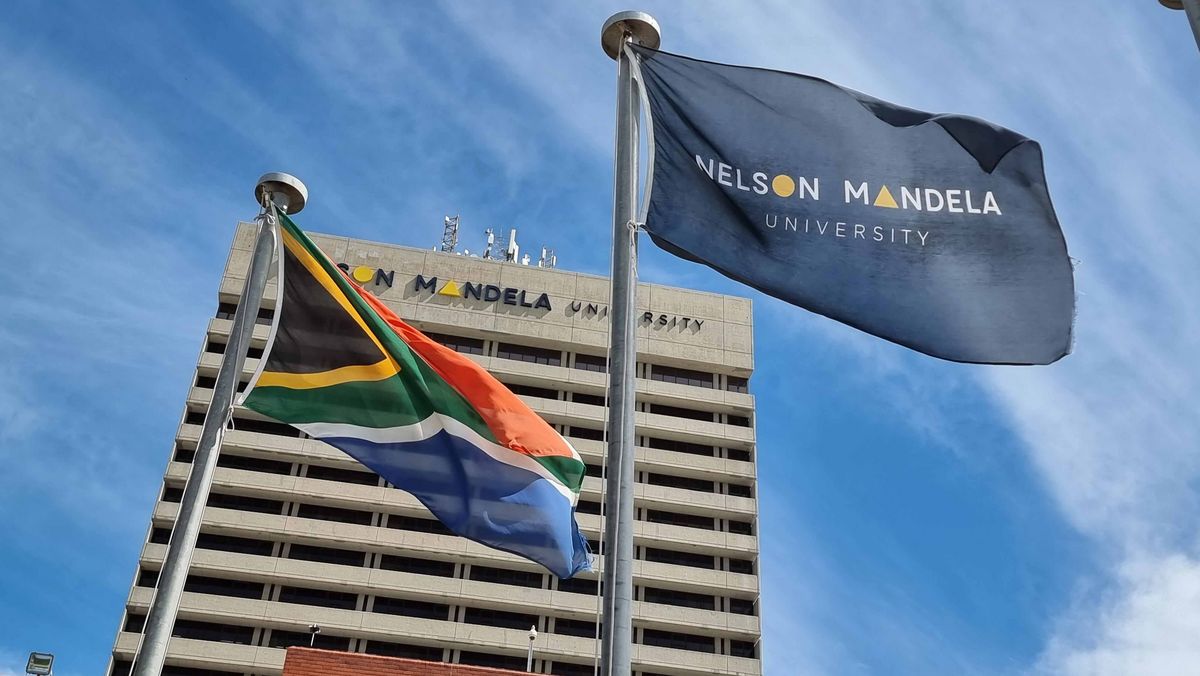 Flaggen der Nelson Mandela University und Südafrikas wehen im Wind vor einem hohen Gebäude.