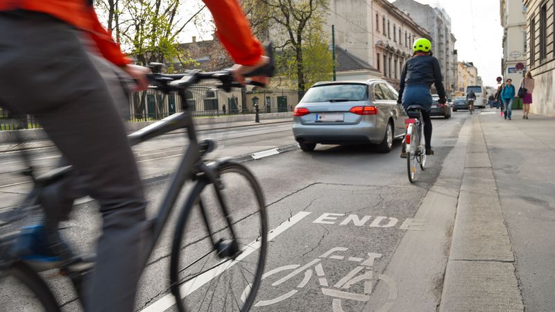 Zwei Radfahrer auf einer engen Straße, ein Auto überholt den vorderen Radfahrer. 