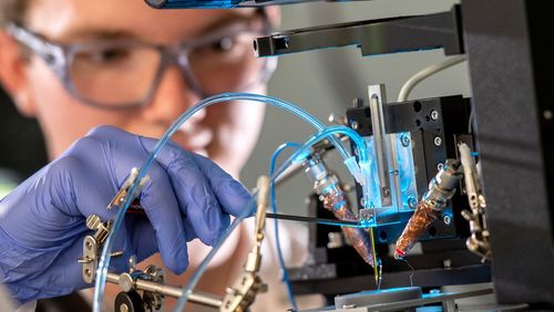 A person wearing gloves makes adjustments to the 3D printer with tubes sticking out of it. 