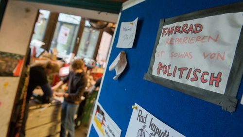 Blick in die Fahrrad-Selbsthilfewerkstatt des AStA. Im HIntergrund sieht man verschwommen zwei Menschen über eine Werkbank gebeugt. Auf der rechten Seite des Bildes ist die blaue Innseite der Eingangstür zu sehen. Daran ist unter anderem ein Schild geklebt, auf dem steht "Fahrrad reparieren ist so was von politisch".