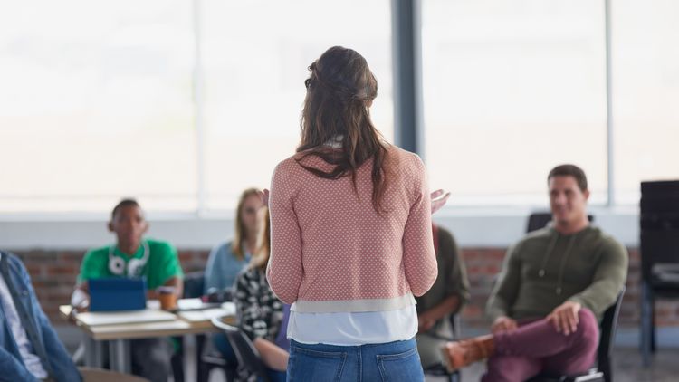 Eine Frau mit rosa Pullover steht vor einer Klasse. 