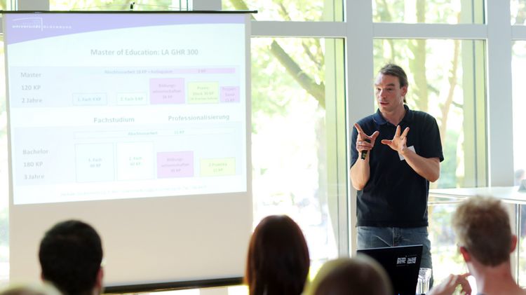 Eine Person hält einen Vortrag mithilfe einer Präsentation vor mehreren Zuschauenden.