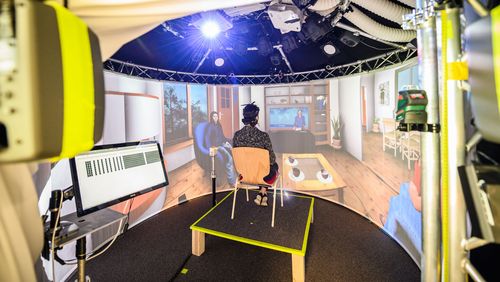 A test person sits on a chair, in the foreground some technical devices (loudspeakers, projectors) can be seen, in the background a living room scene is projected on a screen.