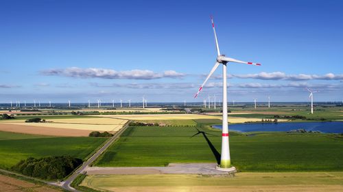 Blick aus größerer Höhe auf die flache, norddeutsche Landschaft aus Feldern, Wiesen und einem See. Im Vordergrund ist eine Windkraftanlage zu sehen, im Hintergrund sind in der Ferne noch zahlreiche weitere Anlagen zu sehen. 