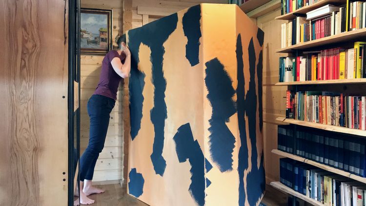 The book stands open in a wooden room, a young woman, who is about the same height as the book, looks inside. The spine of the book is patterned in black and white.