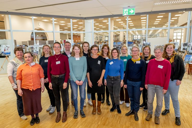 Picture shows a photograph of the participants of the annual network meeting in 2023 in Oldenburg. 15 people are standing in 2 rows in the library of the university and smile at the camera