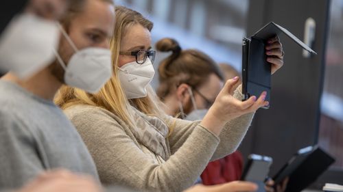 A row of students in a seminar room with masks on their faces, one student holds a tablet in the air.