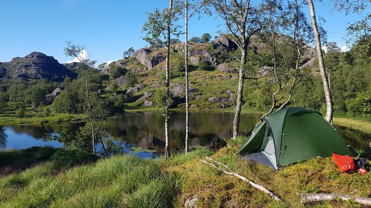 Das Foto zeigt eine norwegische Berglandschaft mit See. Im Vordergrund steht ein Zelt. Im Hintergrund befinden sich bewachsene Berge. Der Himmel ist blau, die Sonne scheint. Die Berge und Pflanzen reflektieren im See.