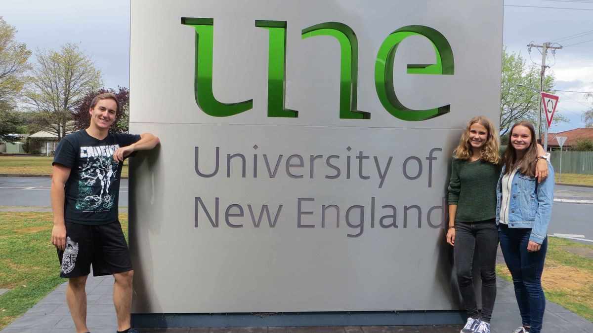 Three young people smile in front of a sign with the UNE logo. Trees and buildings in the background.