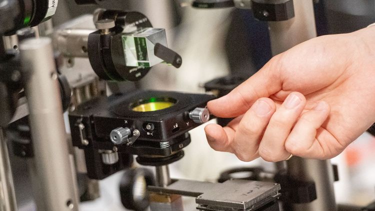 Großaufnahme des Versuchsaufbaus, im Zentrum ein durchsichtiger Glaswürfel, darumherum verschiedene kleine Geräte, die Laserstrahlen umleiten oder reflektieren.