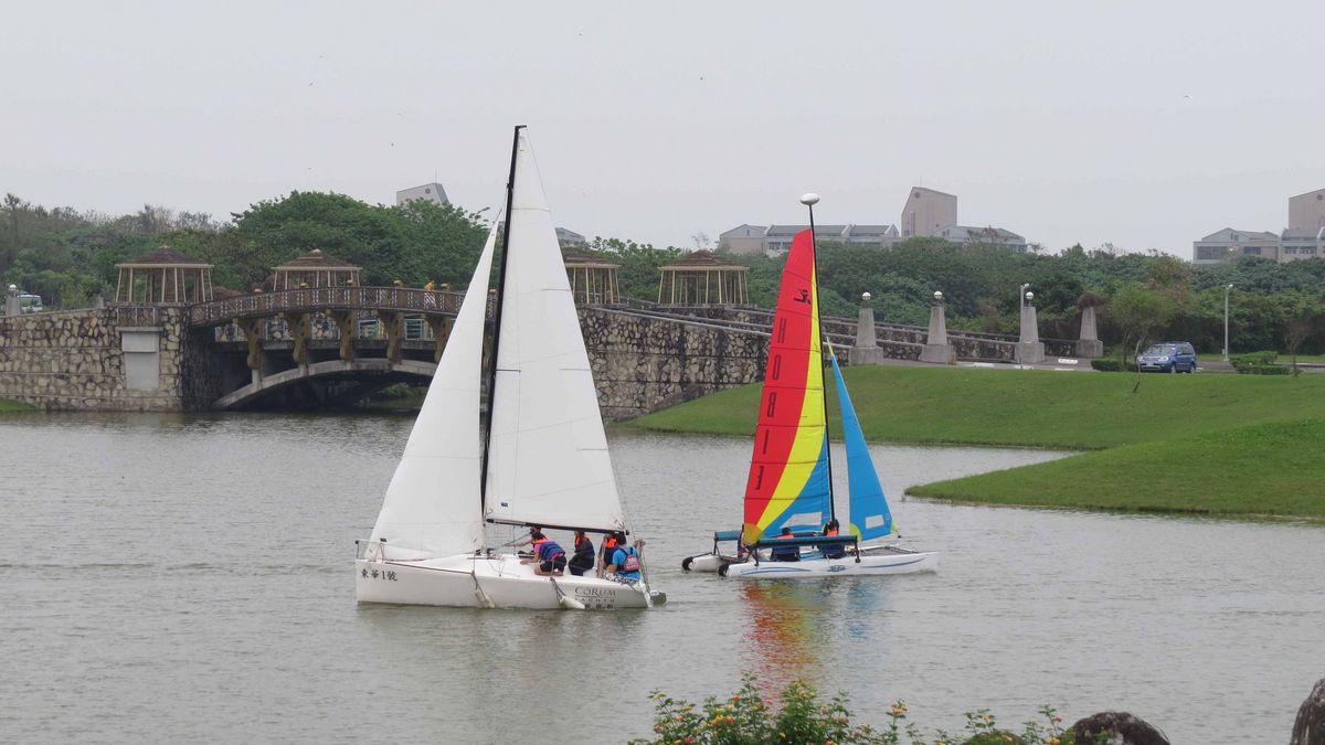 Zwei Segelboote auf einem See vor einer städtischen oder campusartigen Umgebung.