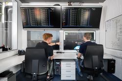 Two people are sitting in a control centre with four big screens.