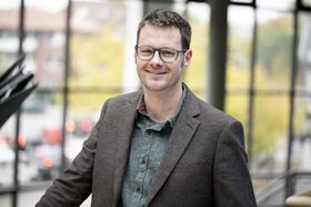 Ein Portrait von Thorsten Balke in seinem Büro: Ein junger Mann mit Brille, Sakko und offenem Hemd vor einem großen Fenster.