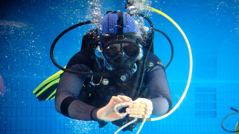 Unterwasserbild. Eine Person im Tauchanzug mit Maske, Sauerstoffflaschen und Flossen verknotet eine dicke Schnur.