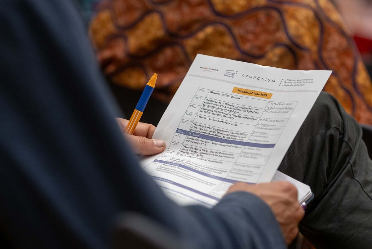 Das Bild zeigt einen Programmplan für ein Symposium, das am 27. Juni 2023 stattfindet. Der Plan wird von den Händen einer Person gehalten und zeigt verschiedene Vorträge und Präsentationen zu Themen wie nachhaltiger Entwicklung, Bildung und den Zielen der Nelson Mandela University.