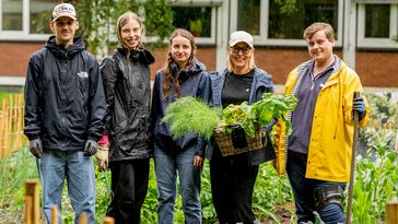 Das Bild zeigt eine Gruppe von fünf Personen, die auf einem Gemüsebeet stehen und in die Kamera lächeln. Zwei Studenten und zwei Studentinnen sowie ihre Professorin tragen Regenjacken, vom Himmel fallen Tropfen. Trotzdem scheinen alle gut gelaunt zu sein. Die Professorin hält einen Korb voller verschiedener Gemüsesorten in den Händen.