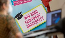 Auf dem Bild sieht man im Hintergrund ein Banner mit dem Schriftzug "Wir sind Fairtrade University". Davor ist verschwimmen ein aufgeklappter Laptop auf einem Stehtisch zu sehen.