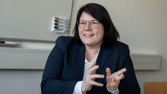 The picture shows Dörte Dannemann. She sits opposite the interviewer and talks. She smiles and gesticulates slightly. A flip chart hangs in the background